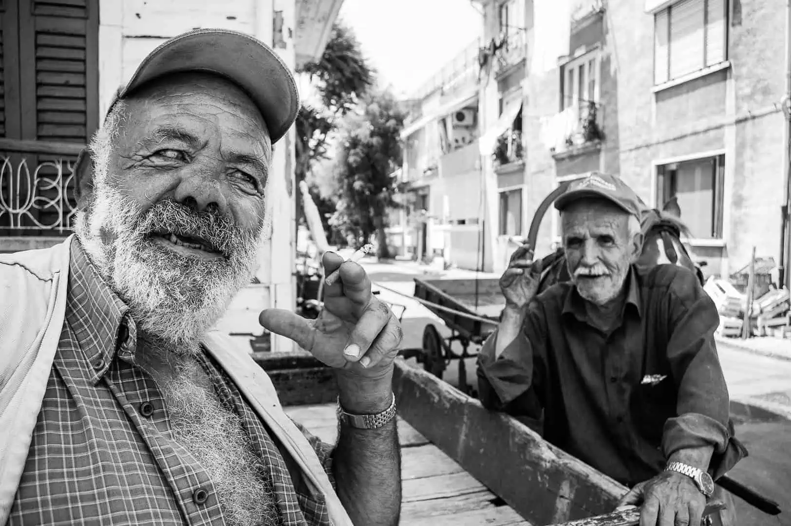 Black and white documentary photograph — Two elderly stablemen sharing a laugh beside a loaded cart while one smokes a cigarette, Büyükada, Istanbul, 2014 by Tarık Kaan Muslu