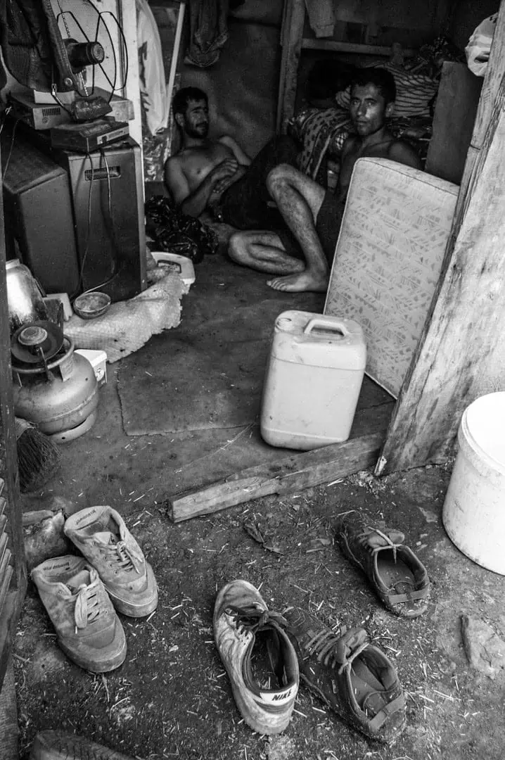 Black and white documentary photograph — Shoes, a water jug, and a television on bare ground inside a stableman's living quarters, Büyükada, Istanbul, 2014 by Tarık Kaan Muslu