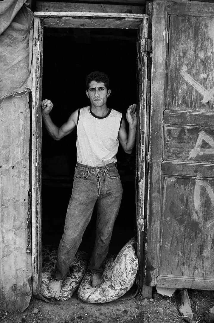 Black and white documentary photograph — Young stableman standing in a doorway between daylight and the dark stable interior, Büyükada, Istanbul, 2014 by Tarık Kaan Muslu