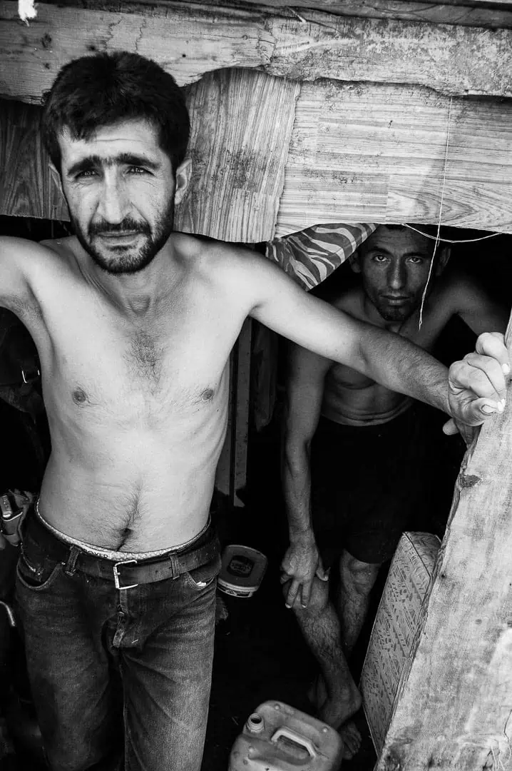 Black and white documentary photograph — Two stablemen standing at the entrance of a makeshift stable shelter, Büyükada, Istanbul, 2014 by Tarık Kaan Muslu