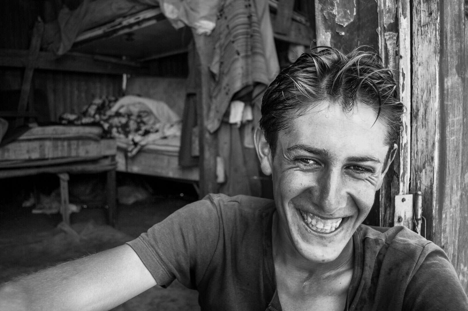 Black and white documentary photograph of a young stableman smiling with a bunk bed and hanging clothes in the background, Büyükada, Istanbul, 2014 by Tarık Kaan Muslu