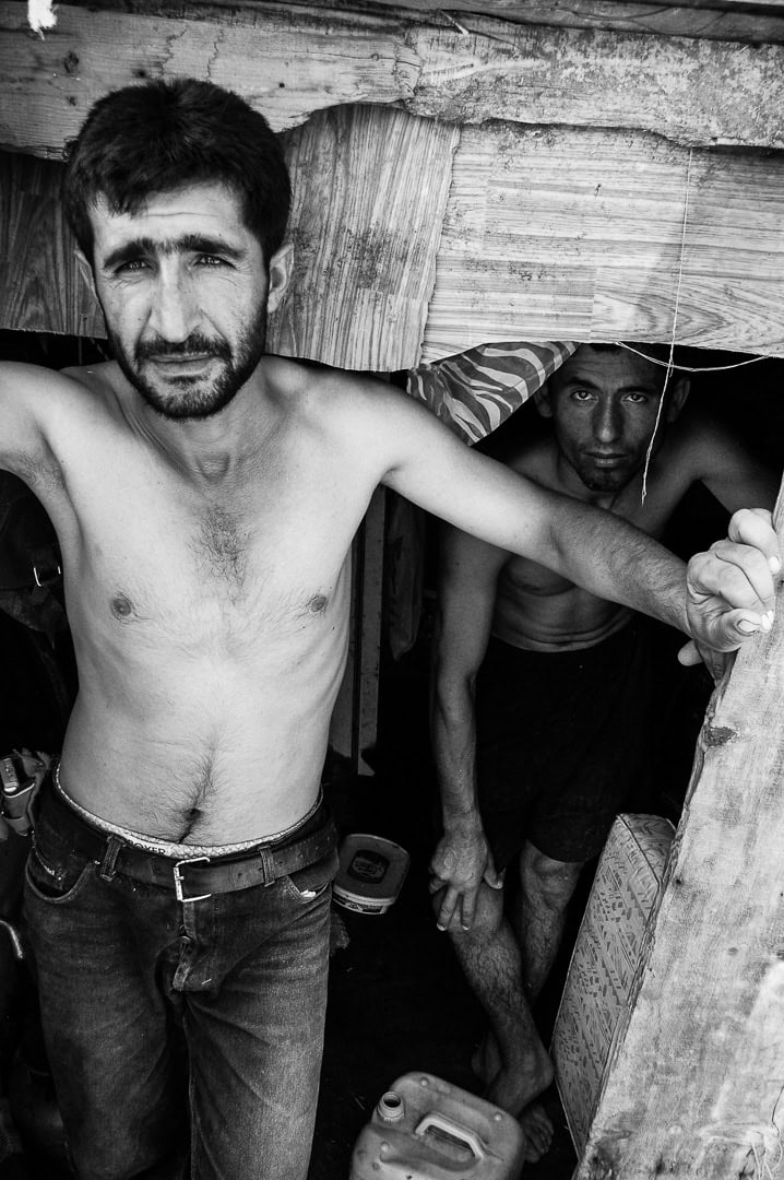 Black and white documentary photograph of two stablemen standing at the entrance of a makeshift stable shelter, Büyükada, Istanbul, 2014 by Tarık Kaan Muslu
